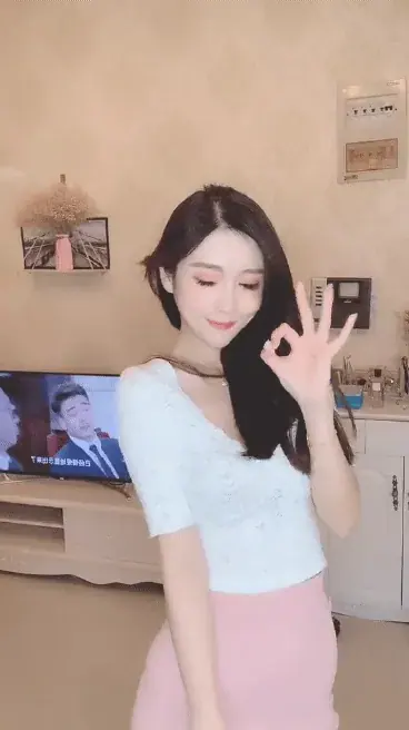 Woman in a white lace top and pink skirt making an OK hand sign in a bright living room with a TV behind her