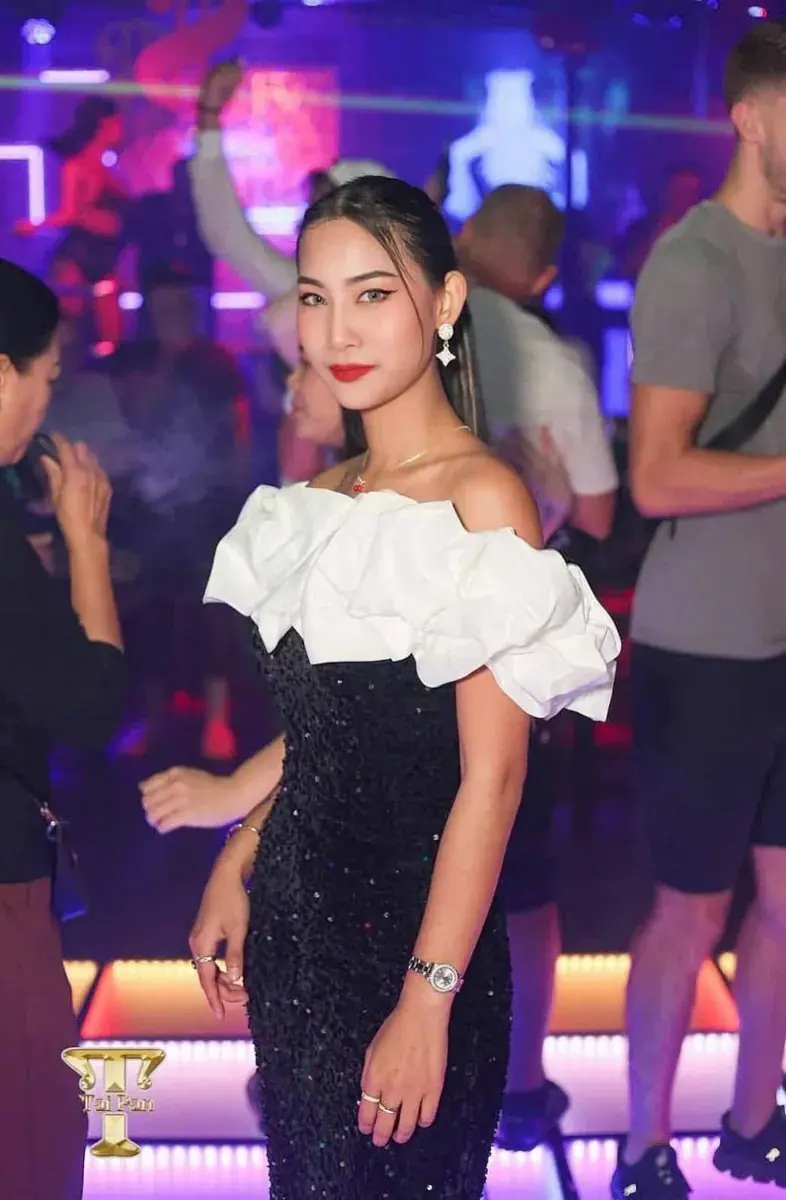A woman in a black sequined dress with white ruffled off-shoulder sleeves poses in a lively nightclub at 69 Bangkok