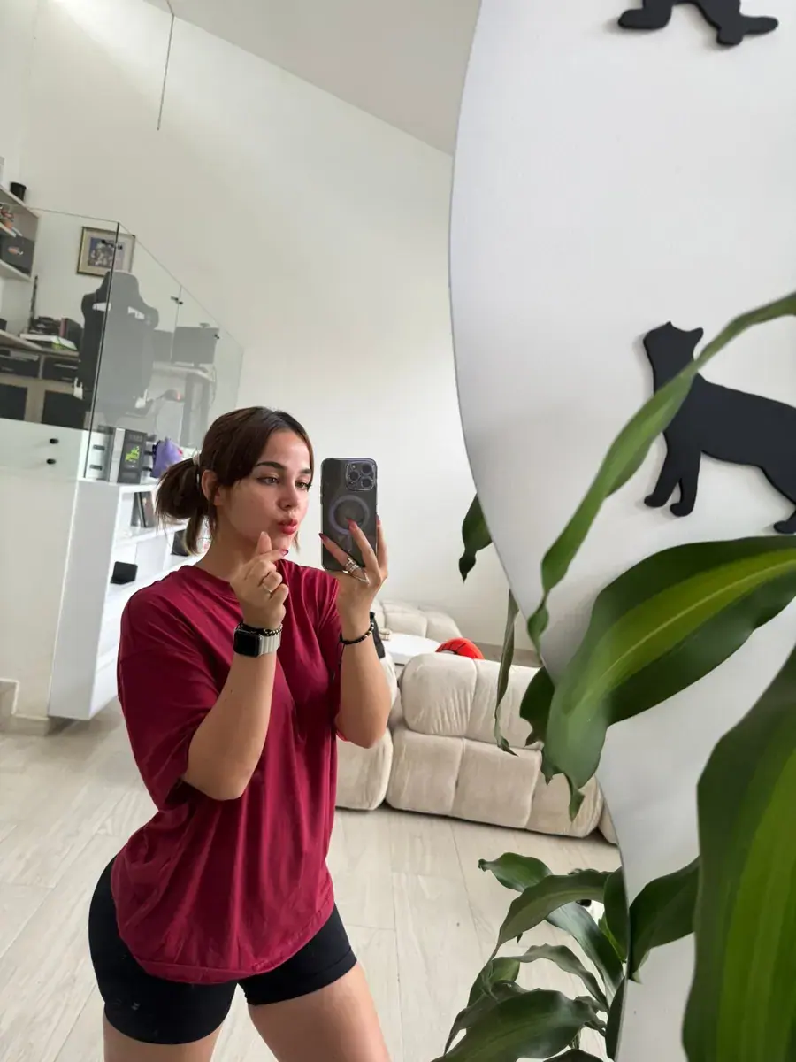 A woman in a red t-shirt and black shorts takes a mirror selfie in a bright living space with a cream sofa and plants
