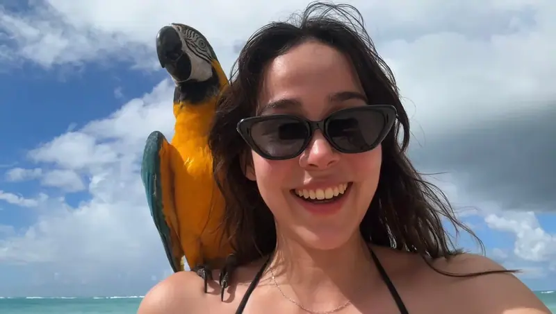 A smiling woman in sunglasses at a sunny beach with a blue and yellow macaw perched on her shoulder
