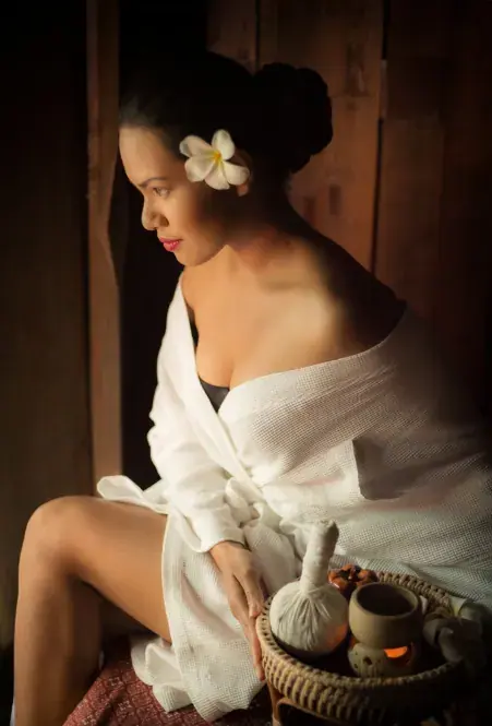 A woman in a white waffle robe with a plumeria flower in her hair sits by a window beside a basket of spa herbal