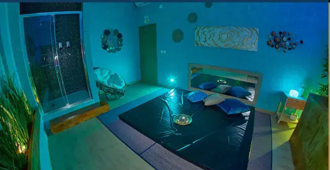 Blue-lit massage room at Artemis SPA in Lisbon with a dark padded mat, cushions, a glass shower and a wicker chair