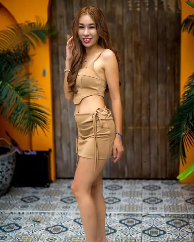 A smiling woman in a tan crop top and matching mini skirt poses by an orange wall at Aya Massage Bangkok