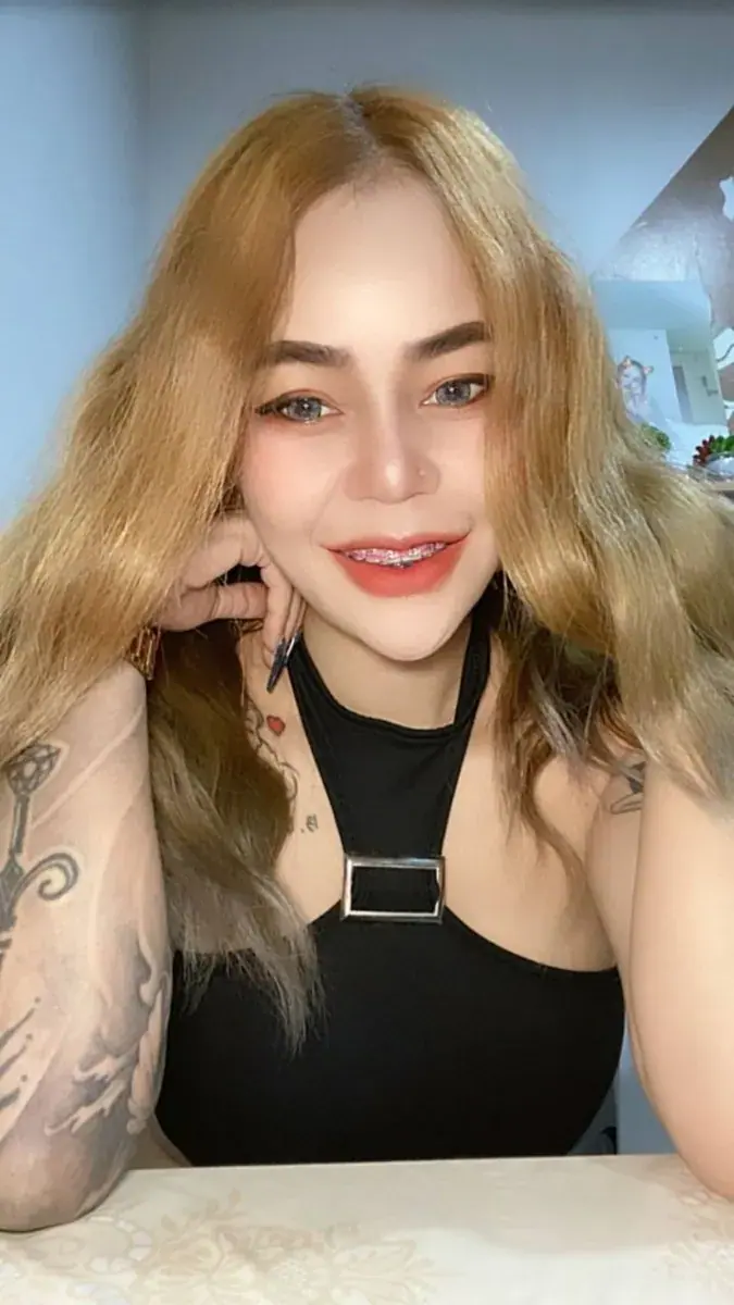 Smiling woman with wavy blonde hair and arm tattoos leaning on a table, in a photo for Bangkok Erotic Massage1 in Bangkok