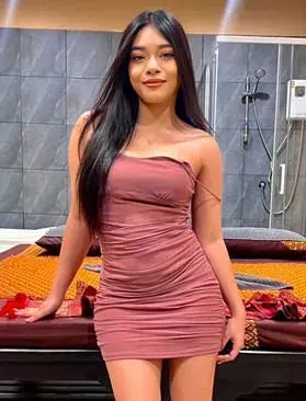Erotic Massage in Thailand: A woman with long dark hair in a fitted dusty-rose dress stands beside a massage bed at Bangkok P