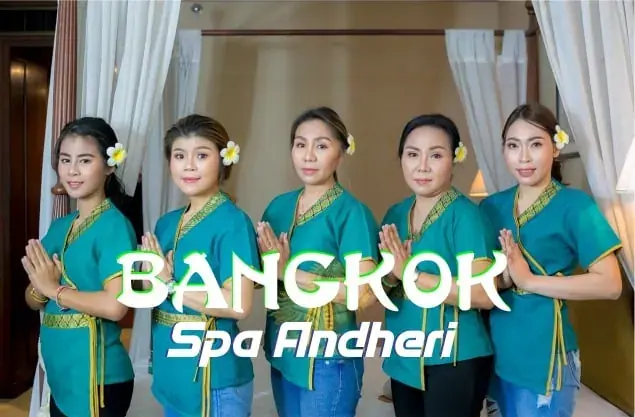 Five women in teal uniforms with flowers in their hair stand with palms pressed together in a draped room at Bangkok Spa