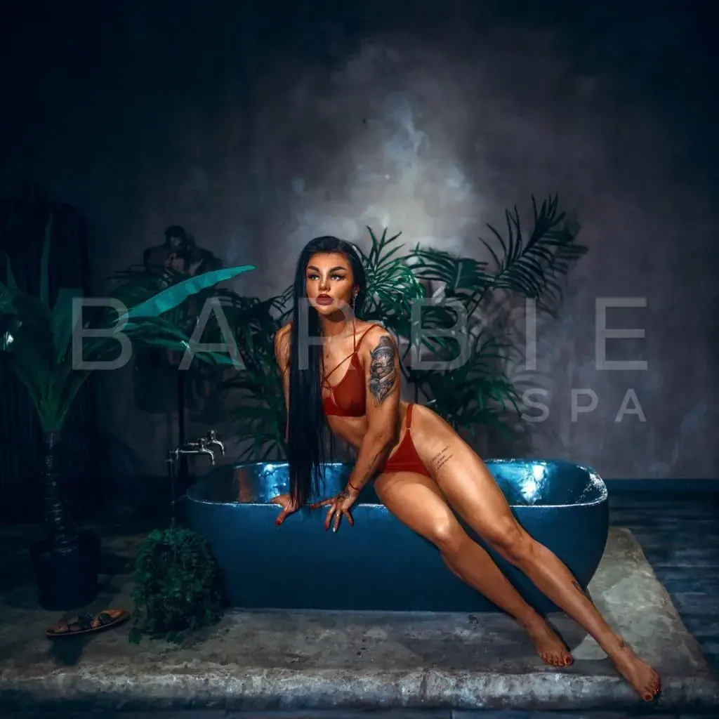 Woman with long dark hair and tattoos posing in a red two-piece by a blue bathtub among tropical plants at Barbi Spa, Moscow