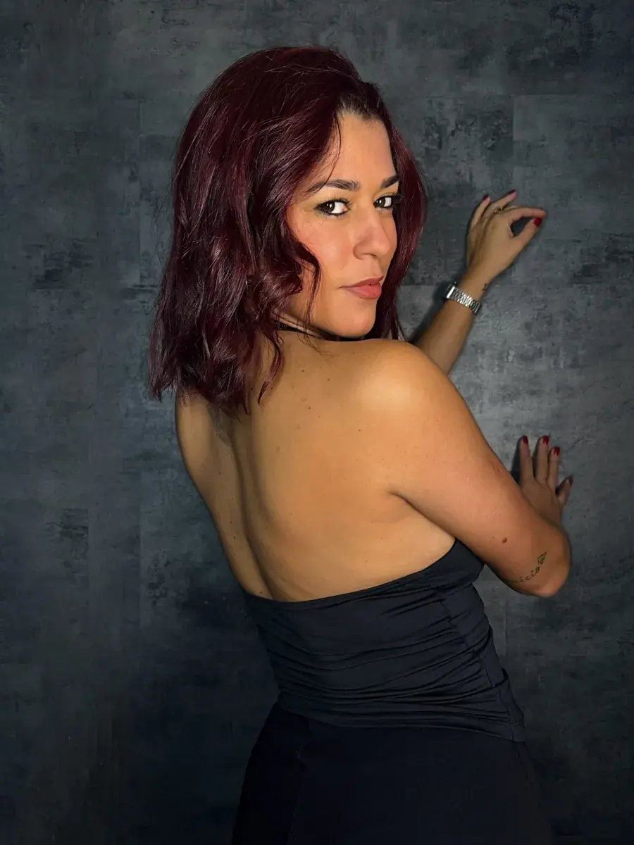 A woman with wine-red hair in a black halter top looks back over her bare shoulder against a dark textured wall at