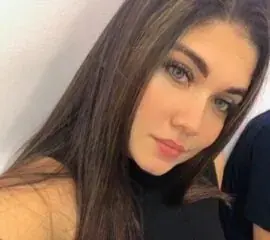 Close-up selfie of a young woman with long brown hair and light eyes, wearing a dark top, indoors