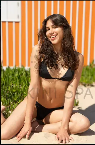 Smiling woman with long dark curly hair sitting on sand in a black bikini, in front of an orange striped wall, for Dakini