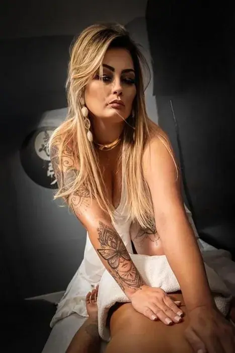 A tattooed blonde masseuse in a white dress leans over a client during a massage at Eros Massagens in Rio de Janeiro