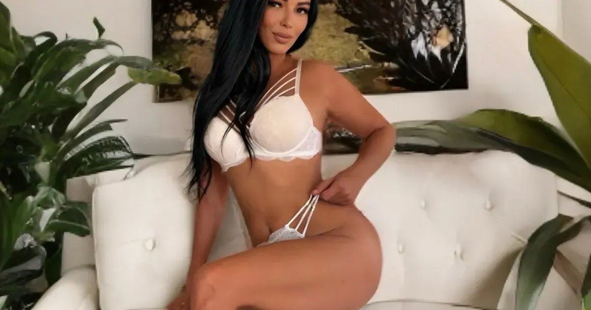 Woman with long dark hair in white lingerie posing on a white tufted sofa among green plants at Erotic Heaven Massage,