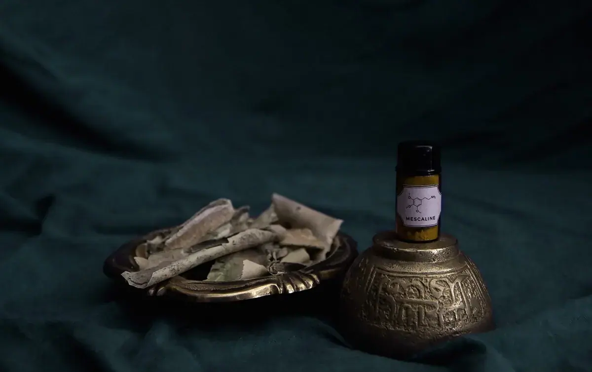 Still life of a brass dish holding dried plant material beside a small labeled vial on an ornate brass base, shown