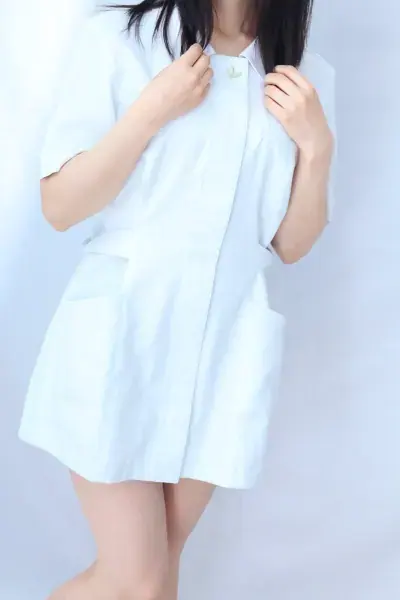 A woman with long dark hair in a white nurse-style uniform poses against a plain light backdrop at Erotic Massage