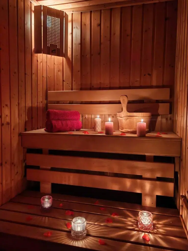 Candlelit wooden sauna at Erotické masáže in Brno, with a red towel, a wooden bucket and scattered rose petals