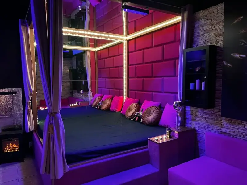 Large platform bed with pink and gold cushions and sheer curtains in a purple-lit room at Erotické masáže, Brno