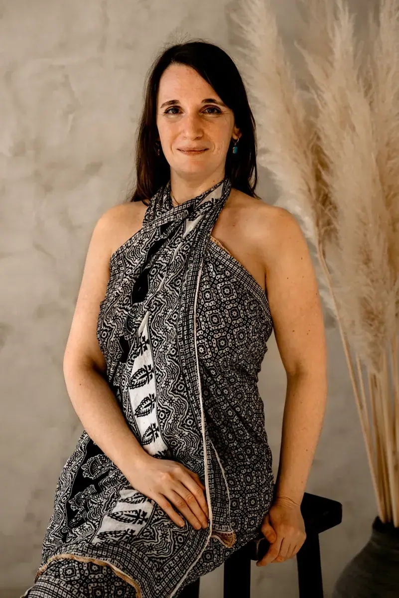 Smiling woman with dark hair in a black and white patterned wrap sitting on a stool beside dried pampas grass at Gaia