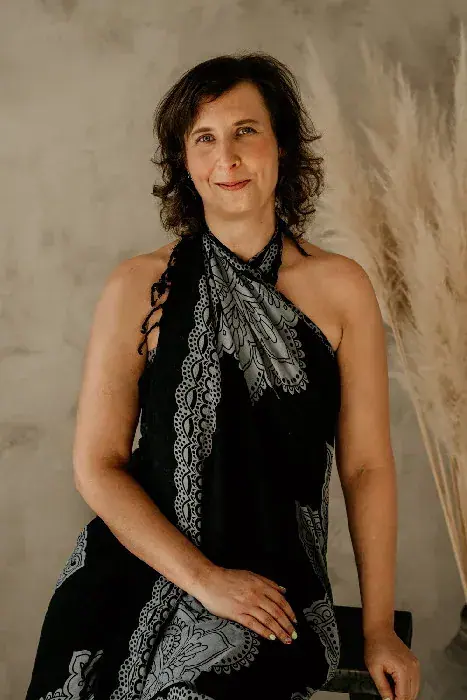 Smiling woman with dark curly hair in a black and grey patterned halter wrap posing beside dried pampas grass at Gaia