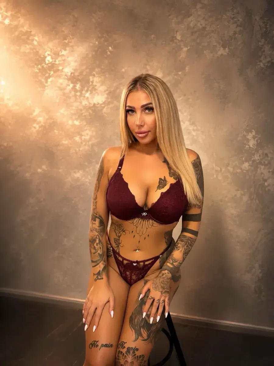 Tattooed blonde woman in burgundy lace lingerie posing against a textured backdrop, listed for Heaven Massage Budapest