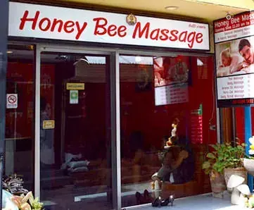 Storefront of Honey Bee Massage in Bangkok with a red sign, glass doors, potted plants and a posted menu board
