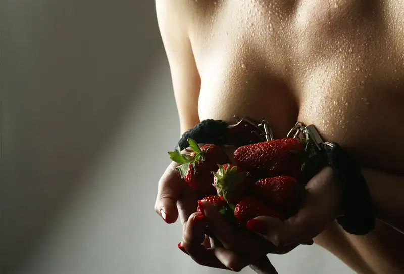 Close-up of a woman's torso with water droplets on her skin, holding fresh strawberries in handcuffed hands, for