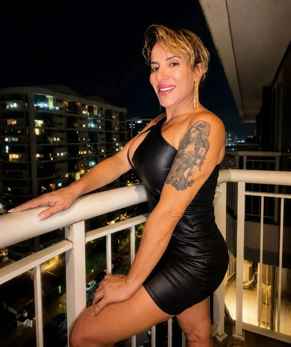 Woman with short blonde hair and an arm tattoo, in a black dress, posing on a night balcony at Lia Gonzalez