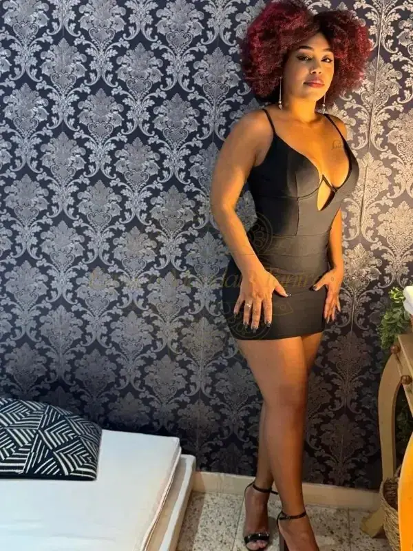 Woman with curly auburn hair in a black dress posing against a damask wallpaper at Mandala Tantra RJ in Rio de Janeiro