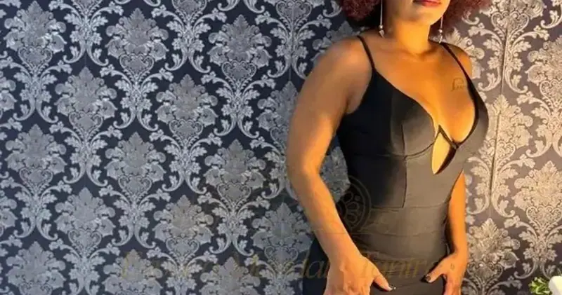 Woman with curly auburn hair in a black fitted dress posing against damask wallpaper at Mandala Tantra RJ in Rio de Janeiro