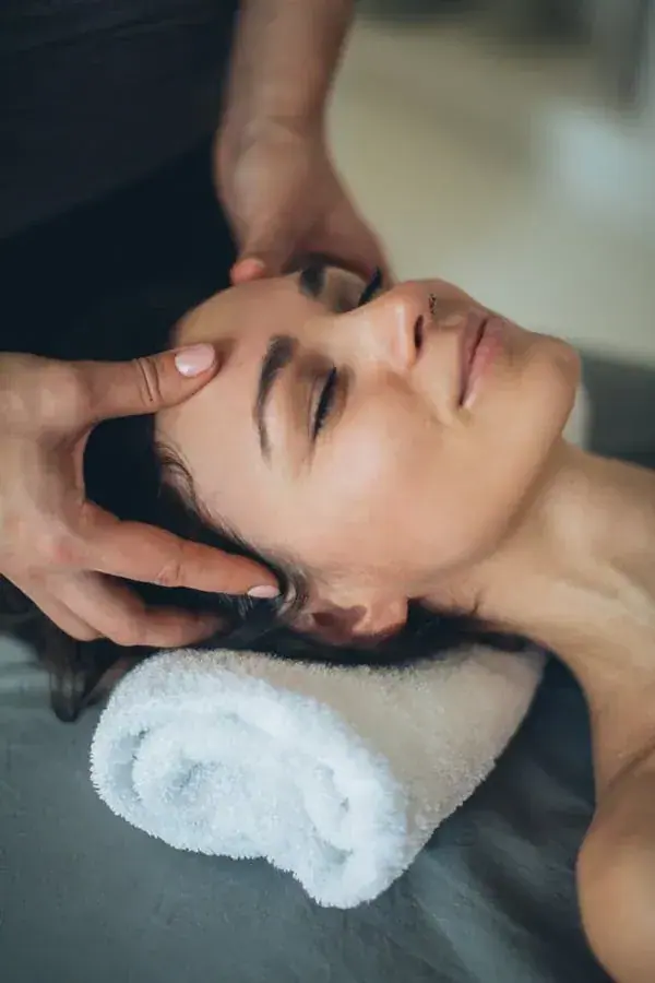 A woman lies with her eyes closed while a therapist massages her forehead, promoting Massage Good in Kyiv