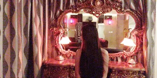 A woman with long dark hair seen from behind, facing an ornate gold mirror in a pink-lit room at Midas Massage, Sydney