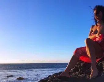 A woman in a red wrap sits on a rocky shore at sunset with the sea and a clear blue sky behind her, for Moment of Touch