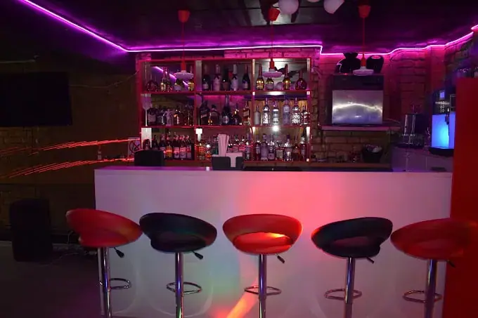 Neon-lit bar at Nadira Masaj in Bucharest, with a white counter, red and black stools and a shelf of bottles