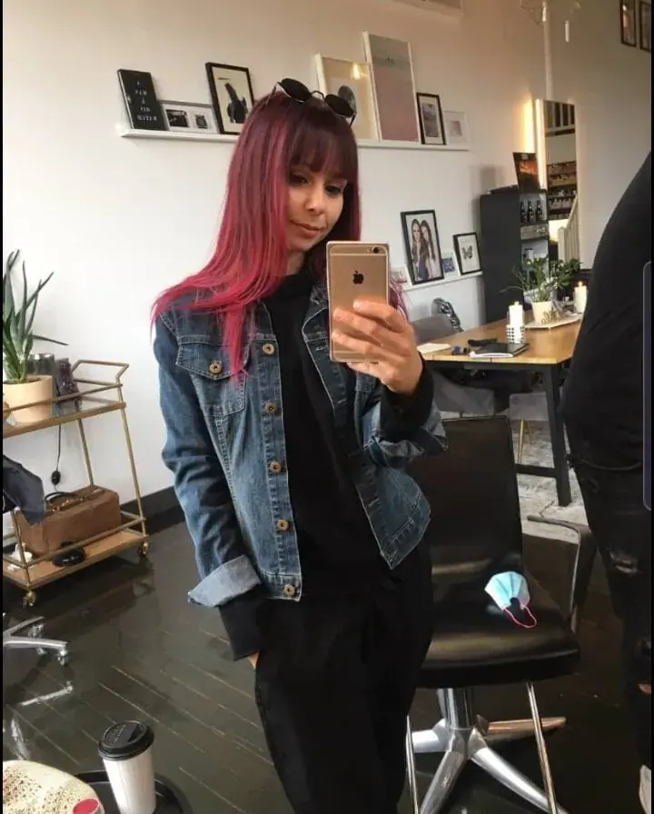 A woman with long red hair and a denim jacket takes a mirror selfie in a salon with a black chair, naming Naomie in Paris