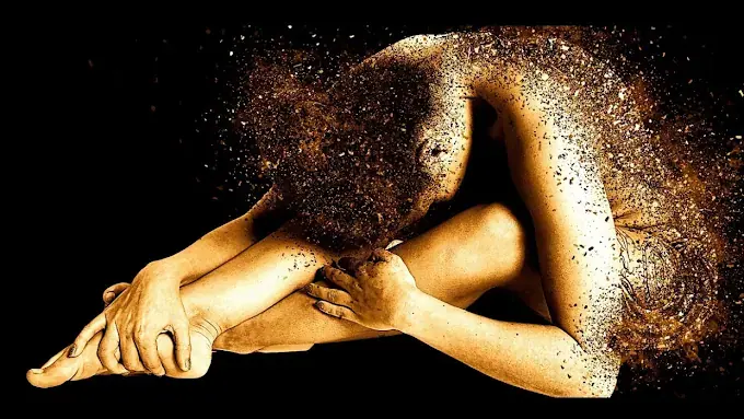 Artistic golden-toned image of a seated figure curled forward, with the head and back dissolving into particles, for