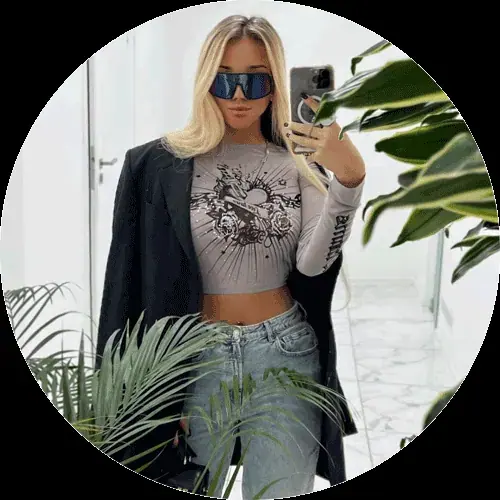 A woman in a gray graphic top, jeans and a black blazer takes a mirror selfie among green plants, used for Nuru Houses in