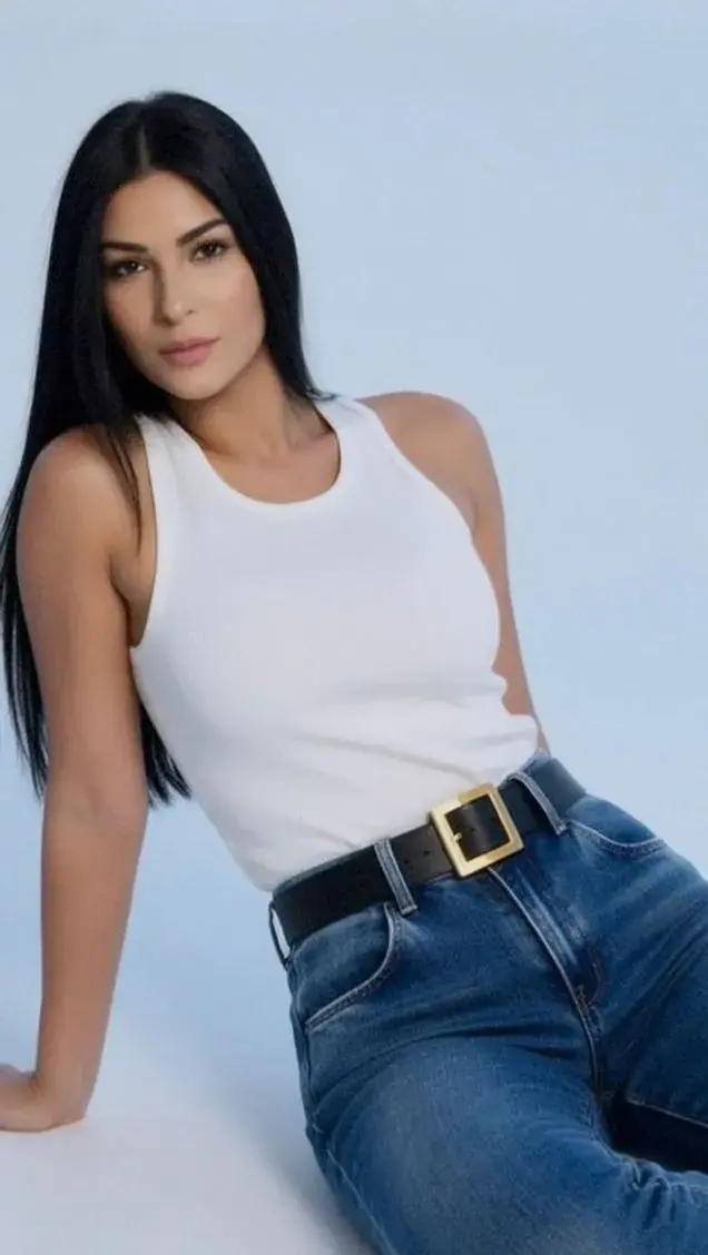 A woman with long dark hair in a white tank top and jeans poses against a pale blue studio backdrop, for Perla Tantric