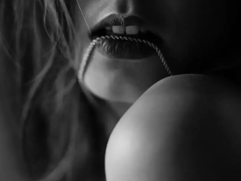 Black-and-white close-up of a woman's lips holding a thin chain, with her shoulder and loose hair in frame, Picasso Bkk,