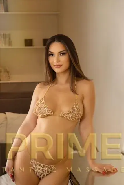 Woman with long brown hair in a gold beaded two-piece posing in a softly lit room at Prime Tantric Massage, London