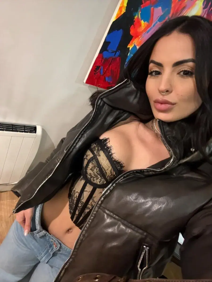 Selfie of a woman with long dark hair in a black leather jacket and lace bustier top, taken indoors near a colorful painting