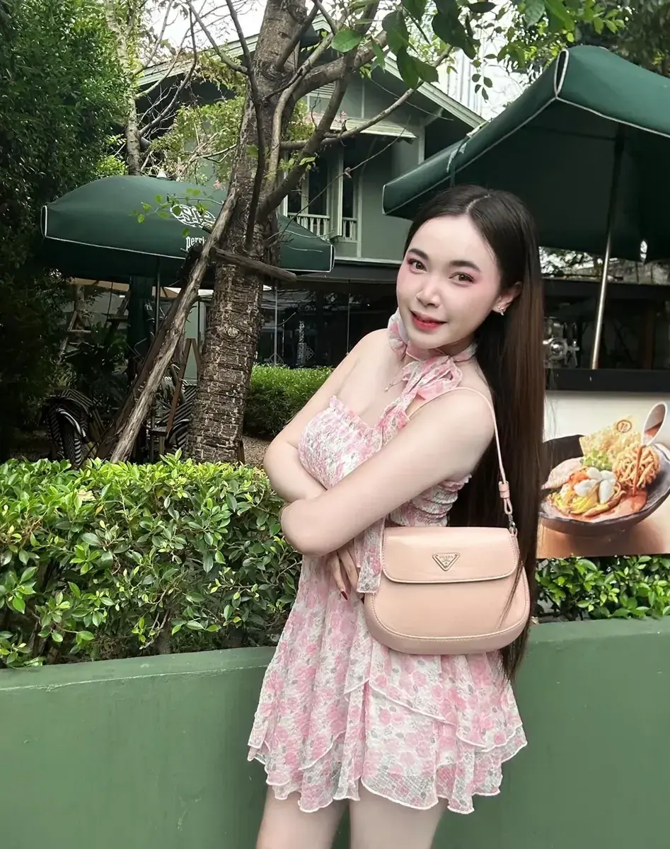 Woman in a pink floral dress with a peach shoulder bag posing outdoors by green hedges and patio umbrellas