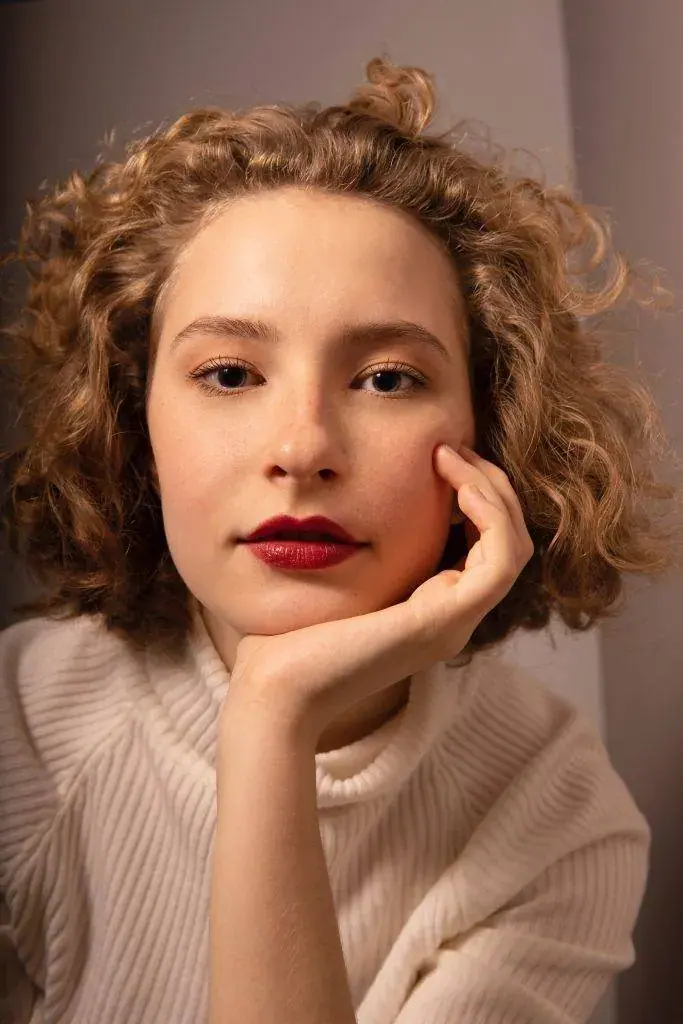 Portrait of a young woman with curly hair and red lipstick resting her cheek on her hand, in a cream sweater