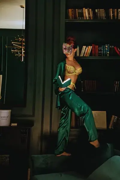 Woman in a green satin outfit and masquerade mask standing on a sofa with a book in a dark library-style room at