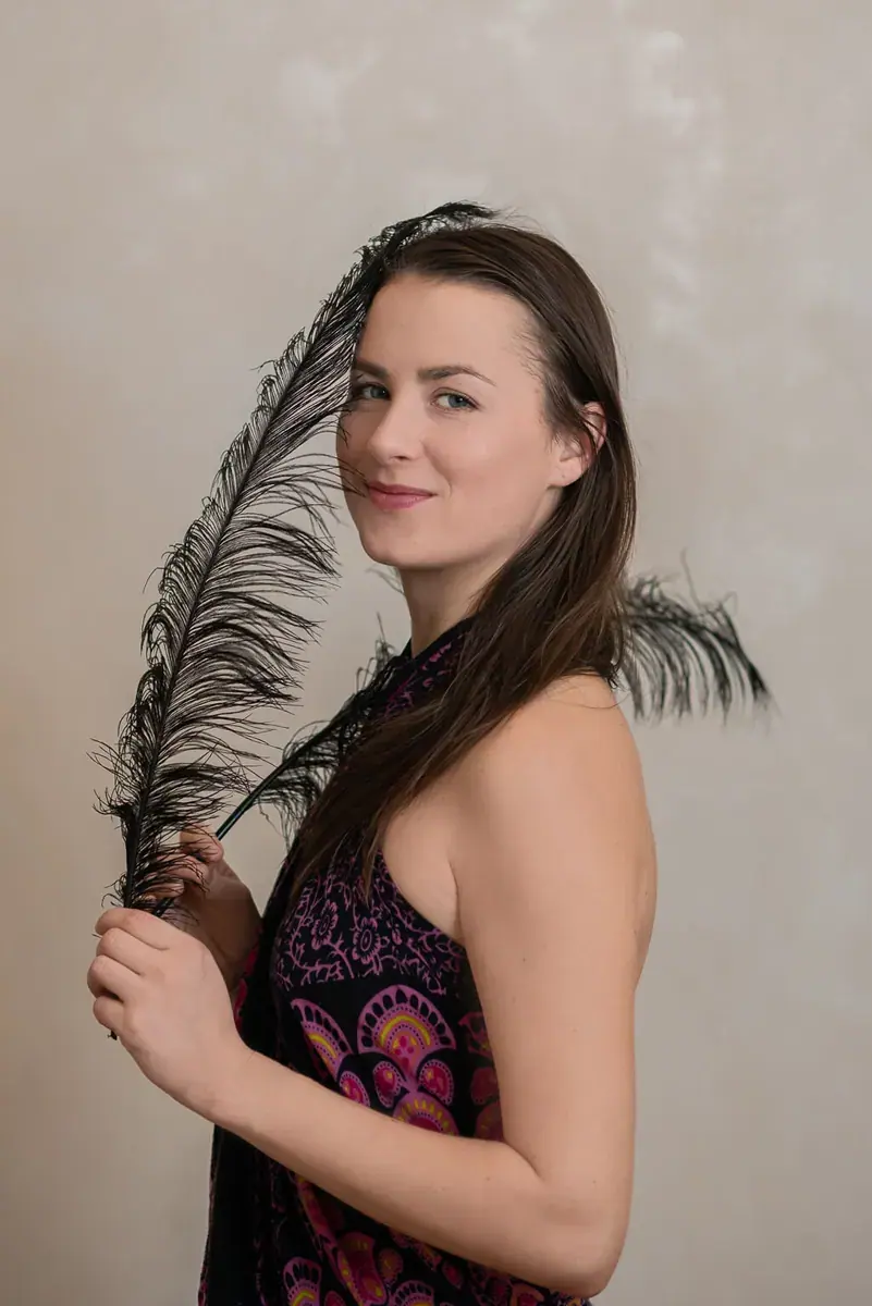 A smiling woman with long brown hair holds a large black feather against a neutral studio wall, Salon Kaira Sayah, Brno