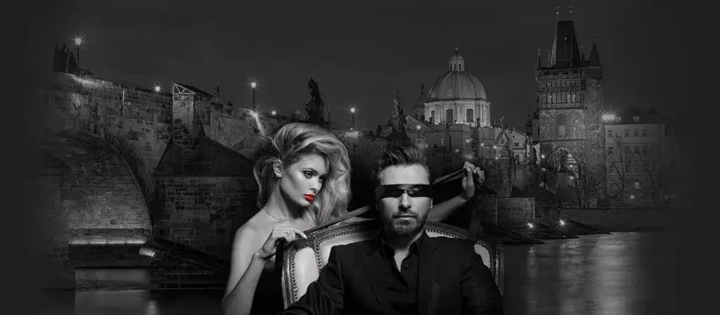 Black-and-white image of a blindfolded man in a suit seated while a blonde woman stands behind him, with a historic