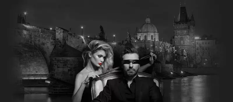 Black-and-white image of a blindfolded man in a suit seated while a blonde woman stands behind him, with a historic