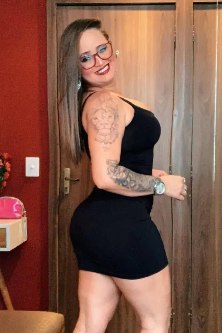 Woman in a fitted black dress and red-framed glasses smiling over her shoulder, Selene Terapias, Rio de Janeiro