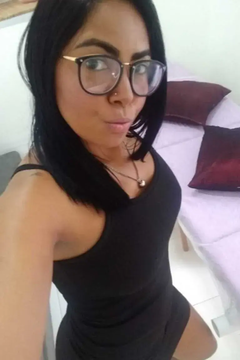 Woman with dark shoulder-length hair and glasses takes a selfie in a black dress beside a massage table at Selene