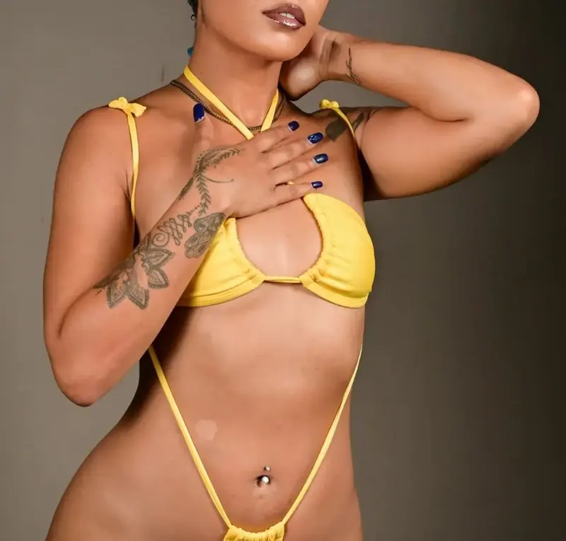 A woman with tattoos and blue nail polish poses in a yellow bikini against a gray studio backdrop, for Spaz Zephyr in