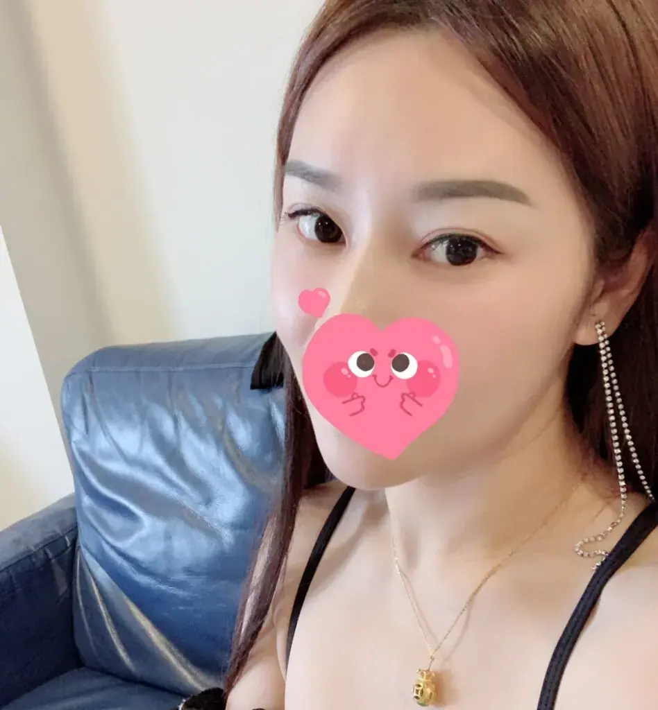 Selfie of a woman with long brown hair and a heart sticker over her face, sitting by a blue leather sofa, for Sydmassage
