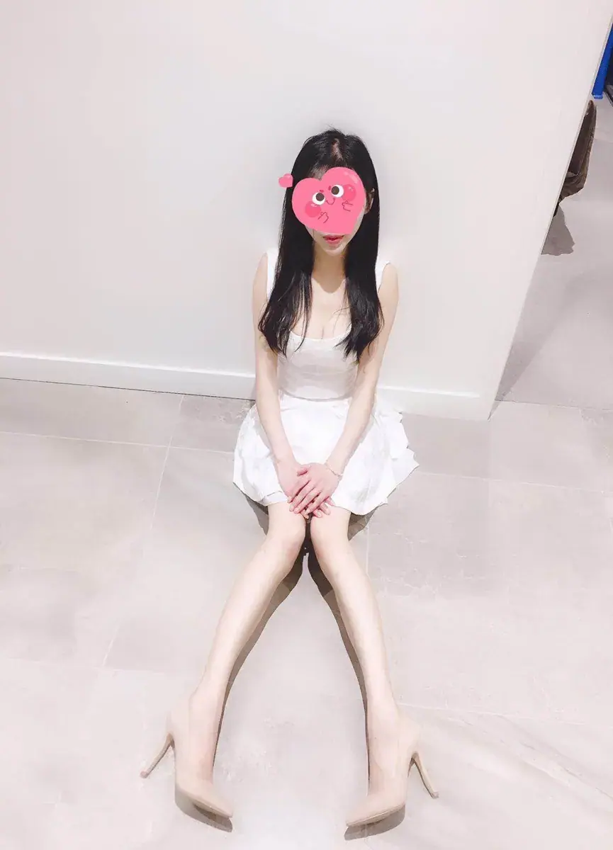 A woman in a white dress and beige heels sits on a tiled floor against a white wall, face covered by a sticker, at